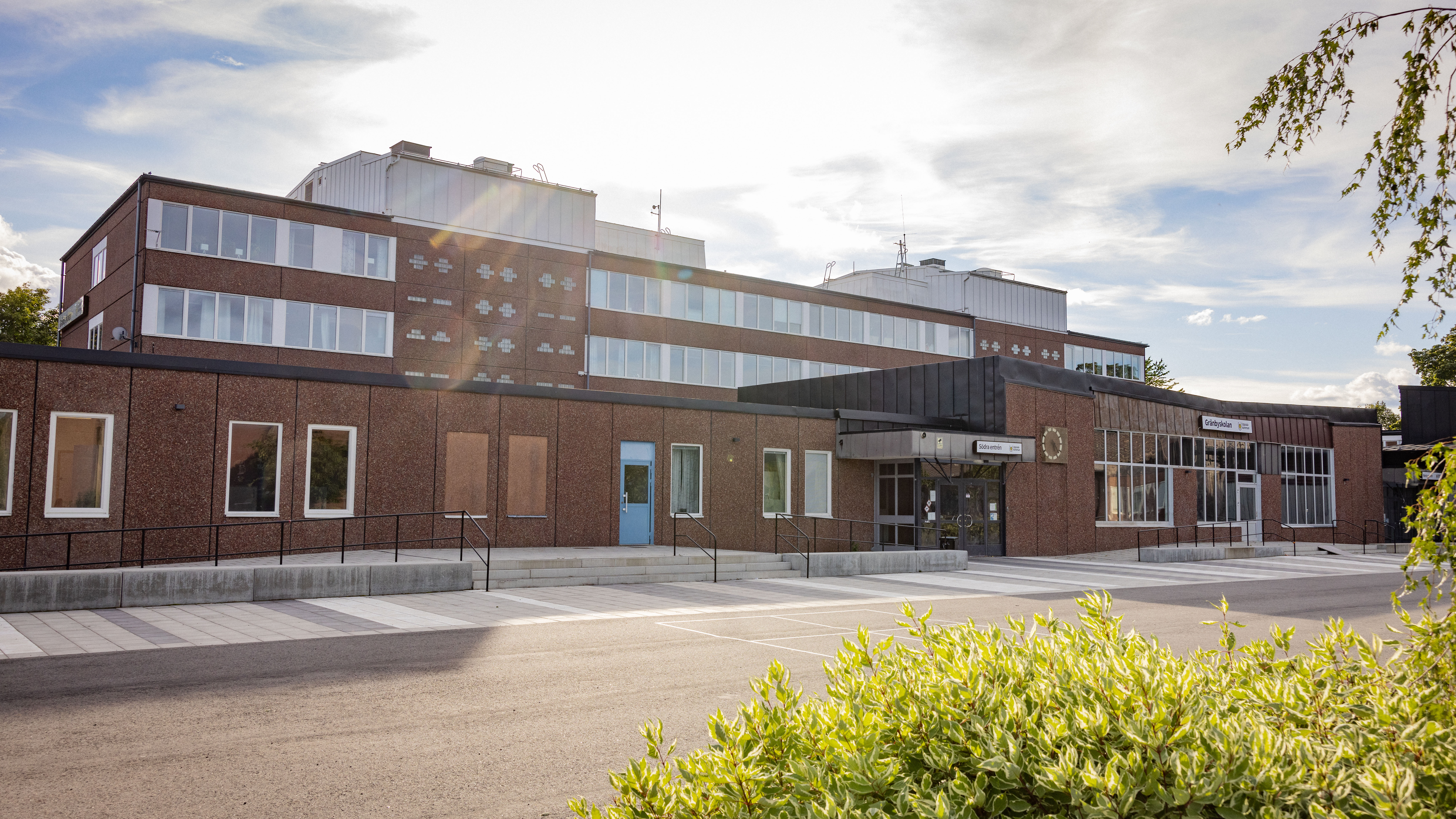 Brun stenskola med gångväg och buskar i förgrunden.