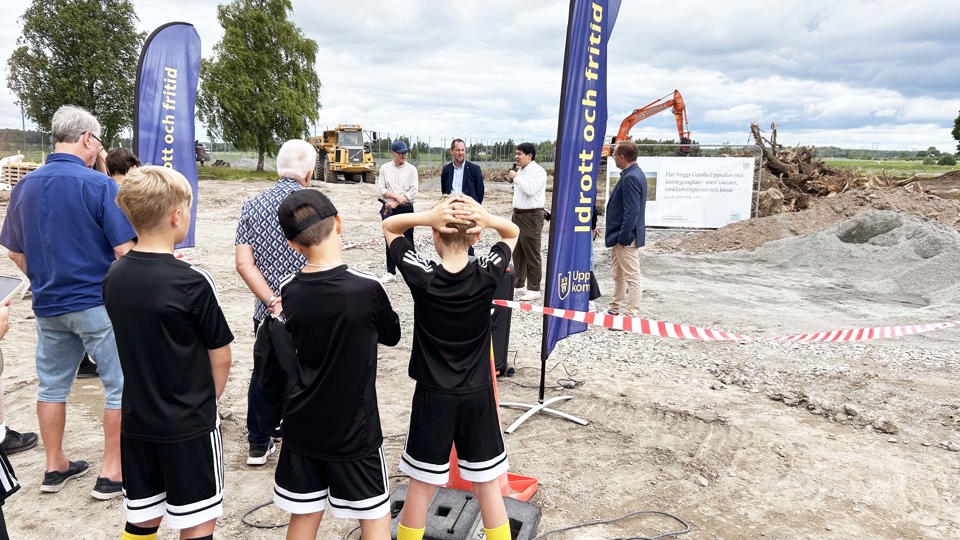 Ungdomar i fotbollskläder lyssnar på politiker som håller tal på en byggplats. 