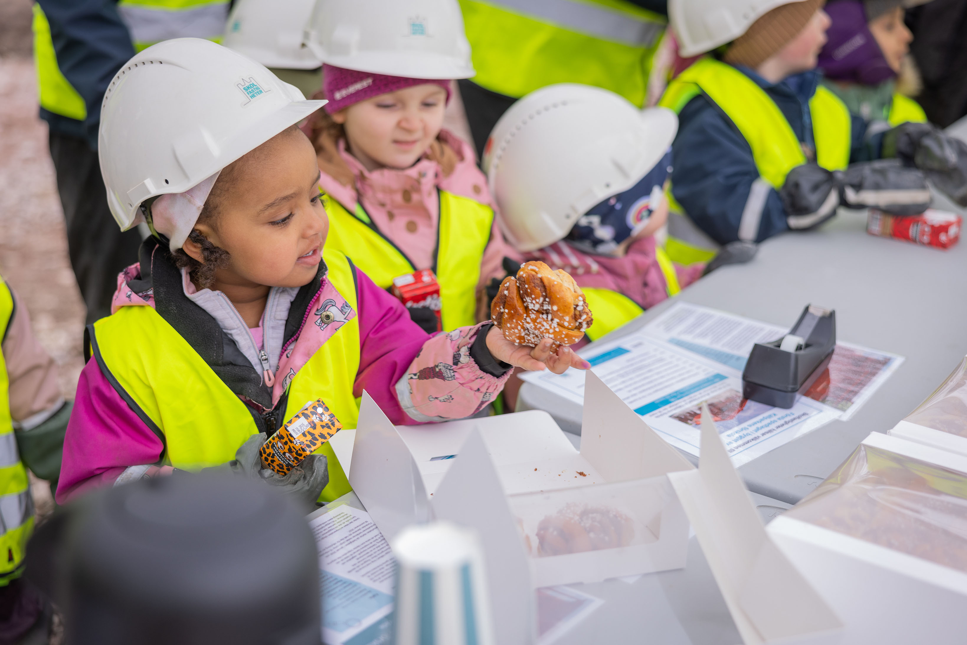 Förskolebarn i bygghjälmar och byggvästar hämtar fika.