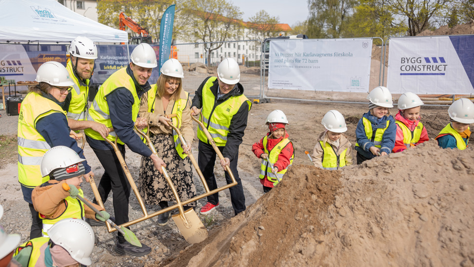 Förskolebarn och vuxna i gula västar och vita bygghjälmar gräver i en sandhög på en byggplats.