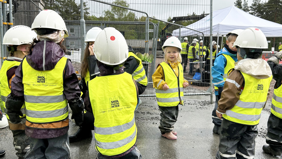 Förskolebarn i gula västar och bygghjälmar besöker en byggplats.