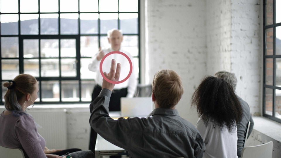 Fem personer runt ett bord, en av personerna räcker upp handen.