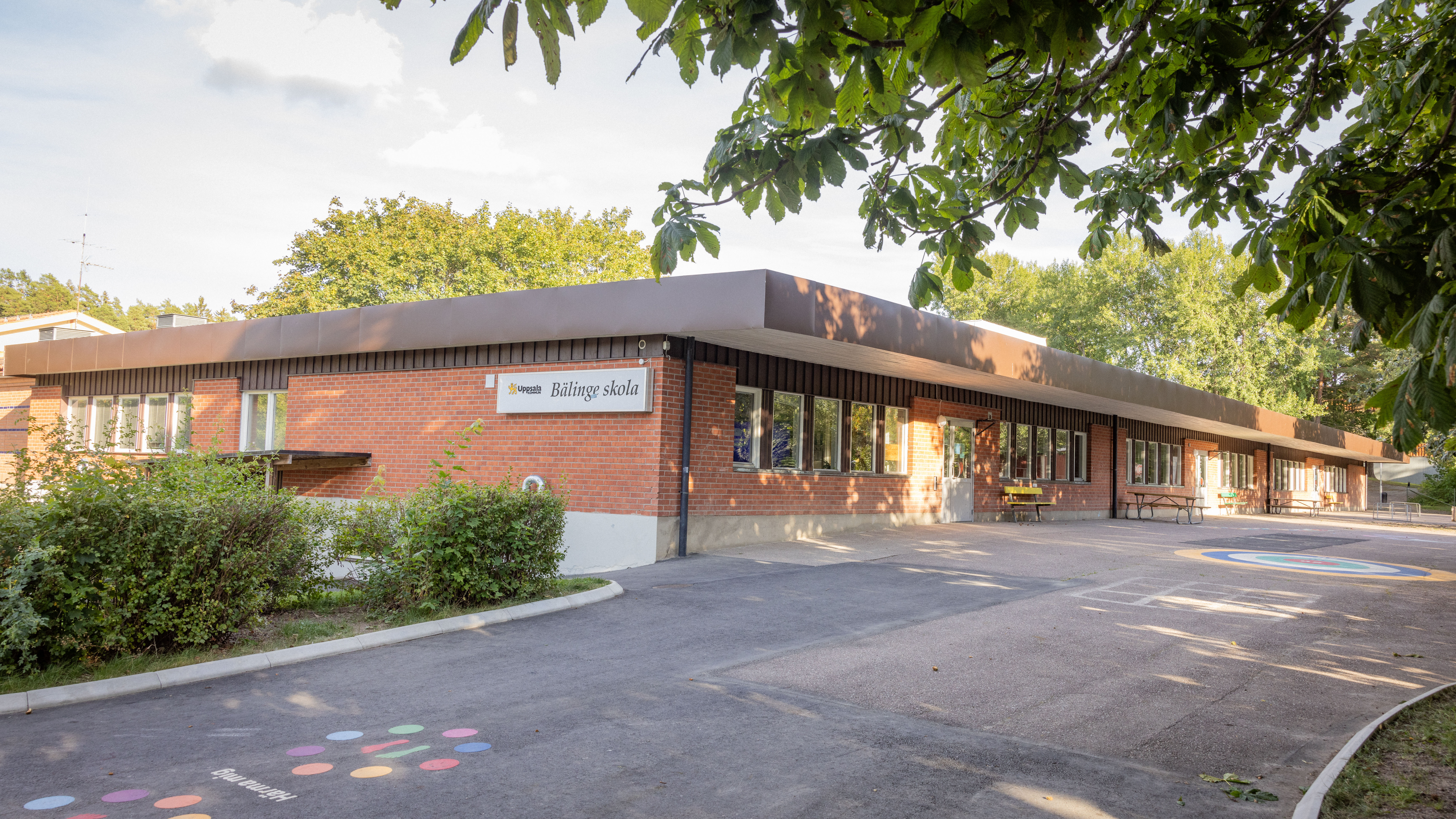 Skolbyggnad i brunt tegel med omgivande gröna träd och buskar